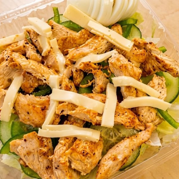 Grilled chicken salad with cheese, cucumbers, tomatoes, and lettuce in a plastic container.
