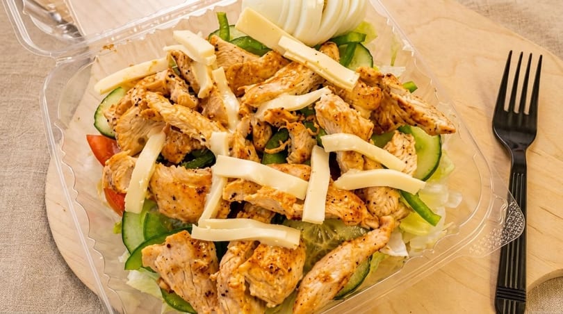 Grilled chicken salad with cheese, cucumbers, tomatoes, and lettuce in a plastic container.